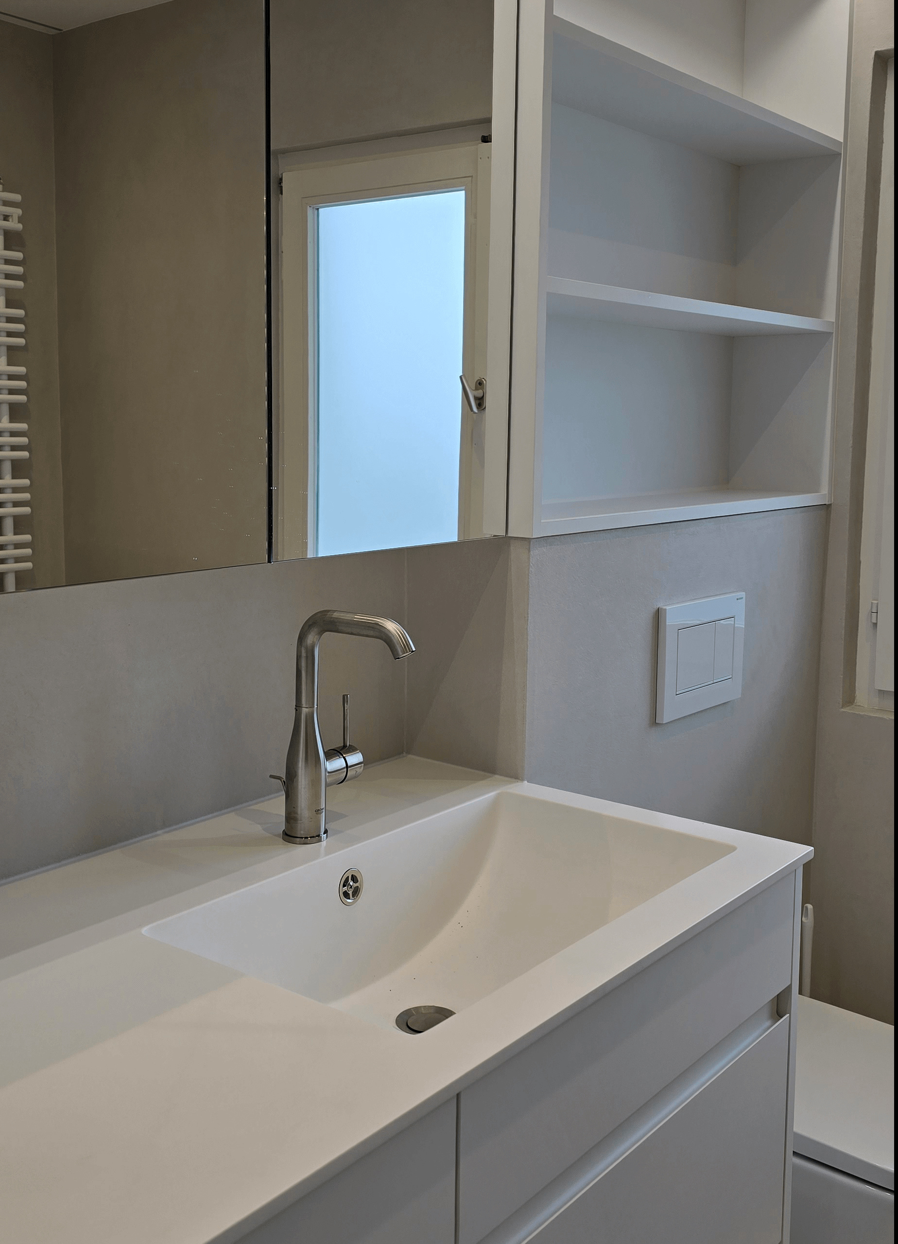 Badezimmer-Moebel in weiss matt, grifflos, weisse Kompaktplatte als Arbeitsplatte mit Spiegelfront und offenem Regal über dem Wc.